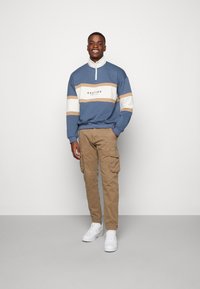 Blaues Sweatshirt mit Viertelreißverschluss, weißen und beigefarbenen Streifen sowie Logo; kombiniert mit beigen Cargo-Hosen und weißen Sneakern. Einfarbiger Hintergrund.