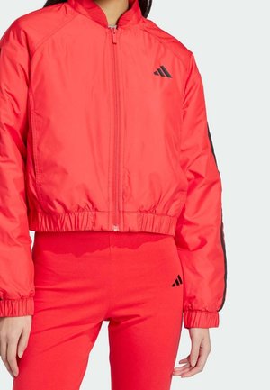 Kvinde iført en skrigrød lynlåstrøje og matchende røde leggings med sorte Adidas-logoer på brystet og låret.