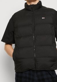 Persona che indossa un gilet imbottito nero senza maniche con una zip sopra una maglietta nera a maniche corte e pantaloni a quadri, con le mani nelle tasche del gilet.