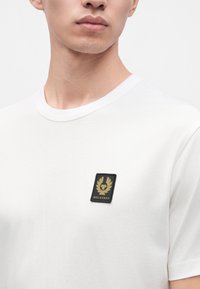 Νέος άνδρας φοράει απλό λευκό T-shirt με μικρό μαύρο και χρυσό λογότυπο Belstaff στο αριστερό στήθος.