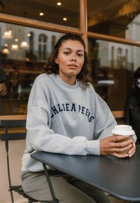 Šedá mikina s námořnickým textem "Chelsea Peeks" na přední straně, texturovaný bavlněný materiál, sedící u černého stolu a držící šálek kávy.