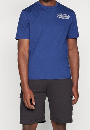 Blue cotton t-shirt with a round neckline and short sleeves, featuring a white Timberland logo on the left chest. Worn with black cargo shorts.