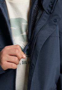 Tamsiai mėlynas viršutinis džemperis su tekstūruotu paviršiumi, užtrauktuku priekyje ir reguliuojamu apykaklės dangteliu; matosi pilkas fliso pamušalas.