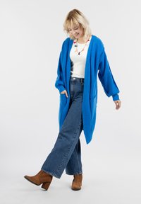 Élénk kék kardigán texturált kötésből, túlméretezett ujjaival és zsebeivel, magas derekú farmerrel és barna sarkú csizmával párosítva.