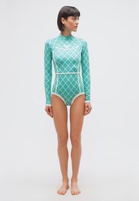 Γυναίκα με κοντά σκούρα μαλλιά που φοράει ολόσωμο μαγιό με μπλε και πράσινο καρό μοτίβο, κάθεται ξυπόλητη μπροστά σε απλό λευκό φόντο.