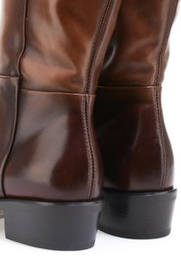 Vue en gros plan de l'arrière de bottes en cuir brun foncé poli avec des talons blocs bas noirs sur fond blanc.
