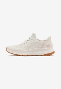 Scarpa da ginnastica beige chiaro con una tomaia testurizzata, dettagli perforati e una suola bianca flessibile. Presenta lacci piatti e una suola in gomma naturale.