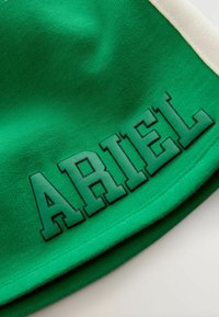 Un tissu vert au toucher lisse présente le mot "ARIEL" en lettres noires en gras et en relief. Le vêtement a un accent de couleur crème.