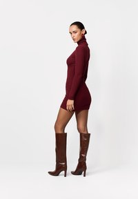 Bordowa mini sukienka z golfem i długimi rękawami, zestawiona z brązowymi, skórzanymi kozakami sięgającymi kolan, z ostrym noskiem i wysokim obcasem.