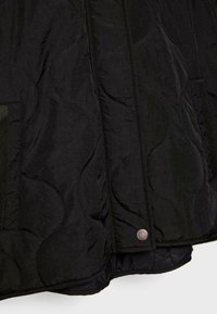 Veste matelassée noire avec un tissu lisse et texturé, des motifs courbés, des coutures proéminentes et un détail de bouton pression à l'ourlet.
