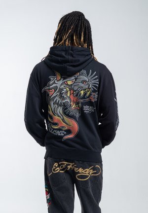 Person med dreadlocks iført en sort hættetrøje med et brølende tigerdesign på bagsiden og sorte jeans med gyldent "Ed Hardy" tekst på lommerne.