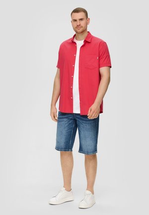 Rotes kurzärmliges Hemd mit Knöpfen und Brusttasche, über einem weißen T-Shirt getragen. Kombiniert mit blauen Denim-Shorts und weißen Sneakers.