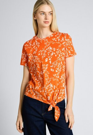 Vrouw draagt een oranje bloemige top met korte mouwen en een knoop aan de zoom, gecombineerd met donkerblauwe spijkerbroek, staand voor een effen achtergrond.