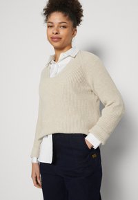 Beżowy sweter z dzianiny z dekoltem w kształcie litery V, o żebrowanej strukturze, noszony na białej koszuli z kołnierzykiem. Model prezentuje luźny krój oraz podwinięte długie rękawy.