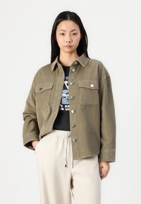 Olive green corduroy jacket with a button-up front, two chest pockets, and a relaxed fit, worn over a graphic black t-shirt and beige pants.