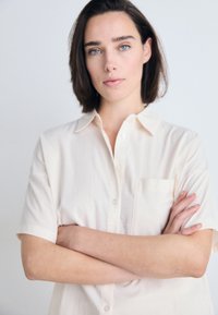 Svetlo béžová krátkoprstá košeľa na gombíky s prednou vreckom, vyrobená z mäkkej látky, s klasickým golierom a voľným strihom.
