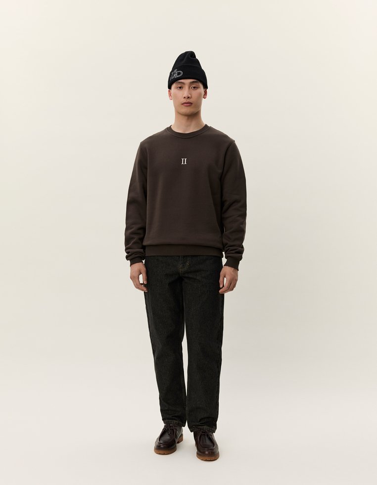 Felpa marrone con un piccolo logo bianco "II", abbinata a jeans in denim neri e un berretto nero. Il modello è in piedi contro uno sfondo neutro.