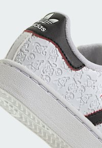 Zapatilla blanca con personajes de dibujos animados en relieve, acento de cuero negro, costuras rojas, suela de goma texturizada y logo de tres franjas en la lengüeta.