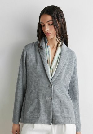 Frau trägt einen strukturierten grauen Blazer mit Knöpfen über einem pastellfarbenen, gemusterten Top, steht vor einer schlichten hellen Wand und blickt nach unten.