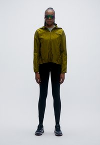 Chaqueta ligera de color verde oliva con capucha y cremallera frontal, llevada sobre leggings negros y zapatillas; la modelo lleva gafas de sol grandes con lentes tintadas.