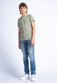 Lichtgroen katoenen t-shirt met kleine tekstdetails, gecombineerd met vervaagde blauwe jeans en witte sneakers. Model staat met de handen langs het lichaam.