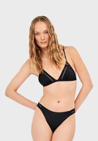 Femme portant un bikini noir avec des détails en dentelle, posant avec une main sur la hanche devant un fond gris clair uni.