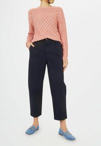 Frau trägt einen rosa strukturierten Pullover, marineblaue, hochgeschnittene, verkürzte Hose und hellblaue Wildleder-Loafer mit goldenen Beschlägen.