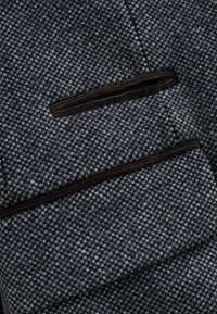 Sötét tengerészkék gyapjú blézer, strukturált, hounds-tooth mintázattal. Fekete cipzáras zsebbel és kontrasztos fekete díszítéssel a zsebeken.