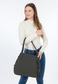 Giovane donna con maglione bianco a coste e jeans blu che tiene una grande borsa nera con dettagli a nappine su sfondo bianco.