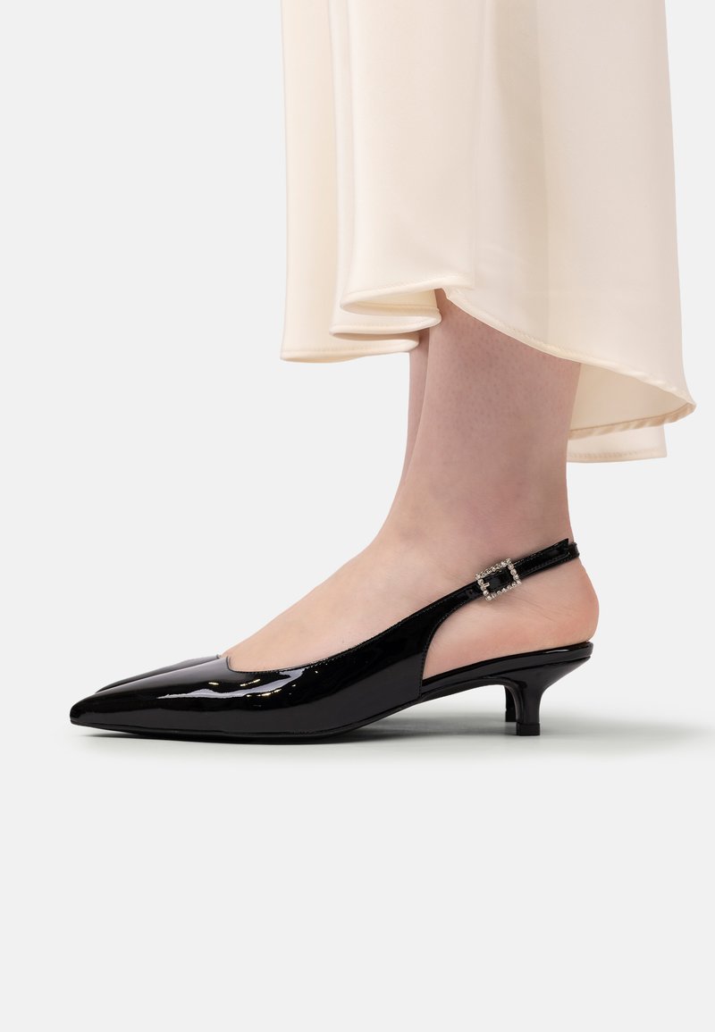 Femeie purtând pantofi slingback din piele patentată neagră cu toc kitten și cataramă cu strasuri, cu tivul unei rochii de culoare crem vizibil.