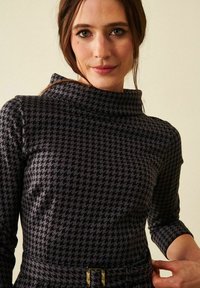 Mekko, jossa on houndstooth-kuvio mustana ja harmaana, funnel-kauluksella, kolmen neljänneksen hihoilla ja vyöllä, jossa on suorakulmainen solki.