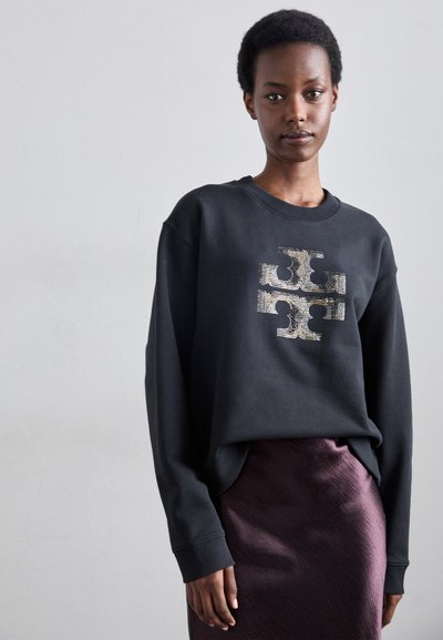 Μαύρη φούτερ μπλούζα με υφασμάτινο σχέδιο στο κέντρο. Διαθέτει στρογγυλή λαιμόκοψη και μακρυά μανίκια. Συνδυασμένη με μια λαμπερή μωβ φούστα.