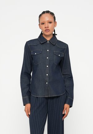 Camisa de mezclilla oscura con cuello puntiagudo, cierre frontal con botones y dos bolsillos en el pecho. Presenta costuras de contraste y un diseño ajustado.