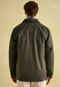 Ciemnozielona pikowana kurtka z kołnierzem z materiału sztruksowego, zapinanymi mankietami i gładką teksturą, pokazana z tyłu na neutralnym tle.