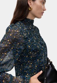 Blomstret bluse i gennemsigtigt sort stof med høj hals, let samlede detaljer og lange puffede ærmer, mens hun holder en sort taske.