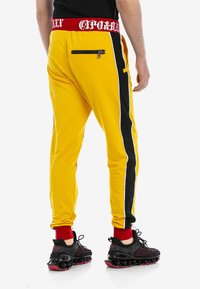 Gelbe Jogginghose mit einem schwarzen Seitestreifen, ausgestattet mit einem roten elastischen Bund mit weißem Text und einer Reißverschluss-Tasche hinten. Schwarze Sneakers mit roten Akzenten.