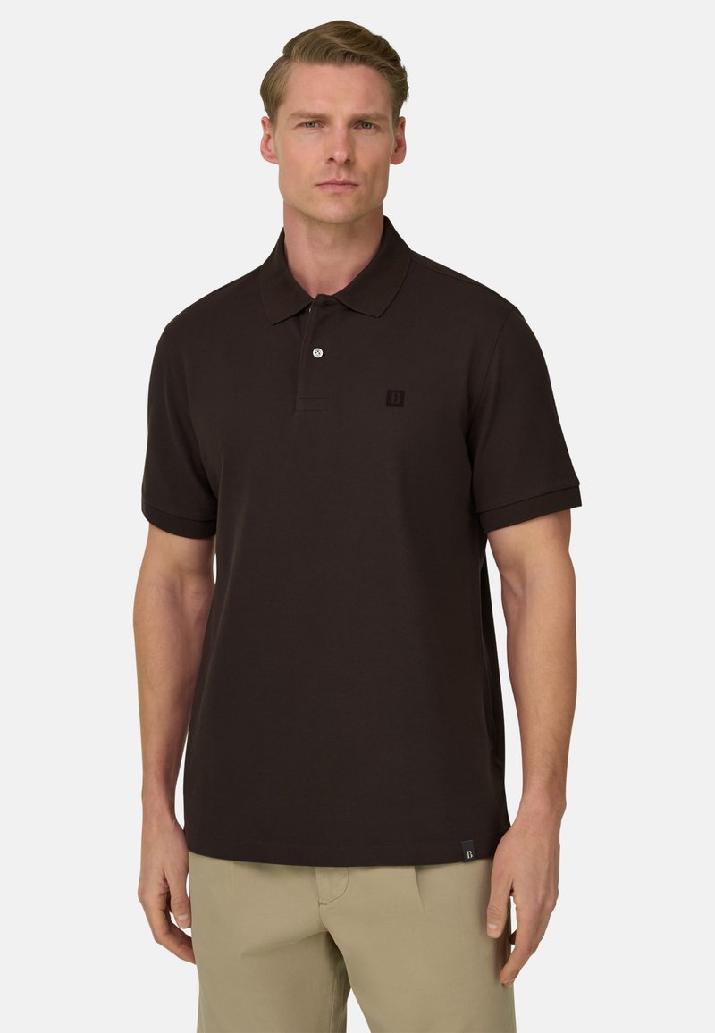 Homme portant un polo noir à manches courtes et un pantalon beige, debout devant un fond clair uni.