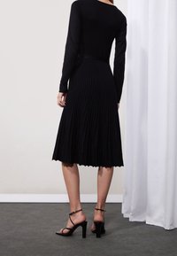 Femme portant un haut à manches longues noir et une jupe midi plissée, avec des sandales noires à talons hauts, debout sur un sol sombre près de rideaux blancs.