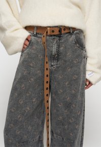 Jeans grises con un estampado sutil y un cinturón marrón. Los jeans presentan una impresión de logo discreta y un ajuste relajado; el cinturón tiene herrajes negros y un acabado texturizado.