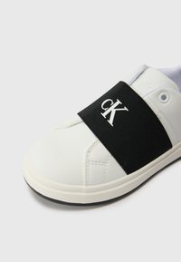 Hvit sneaker med en svart elastisk stropp med "ck"-logo. Glatt lærtekstur, rund tå og gummisåle. Minimalistisk design.