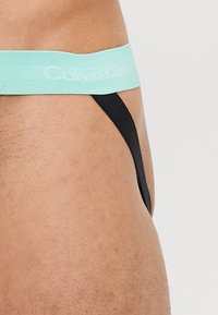 Crne tange s elastičnim pojasom svijetloplave boje marke Calvin Klein, s glatkom teksturom i minimalističkim dizajnom, prikazane sa strane.