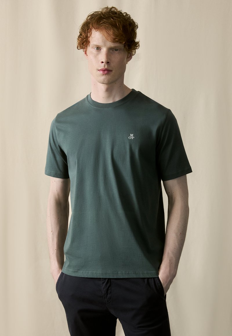 T-shirt en coton vert foncé avec un col rond, des manches courtes et un petit logo blanc sur la poitrine gauche. Texture lisse, coupe décontractée.