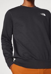 Sudadera negra de manga larga, con puños y dobladillo acanalados. Presenta un pequeño logotipo blanco en la parte superior izquierda del pecho, con una textura de tela suave.