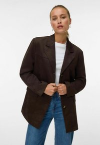 Blazer in suede marrone con rever a punta, due bottoni e tasche frontali. Abbinato a una camicia bianca e jeans in denim blu.