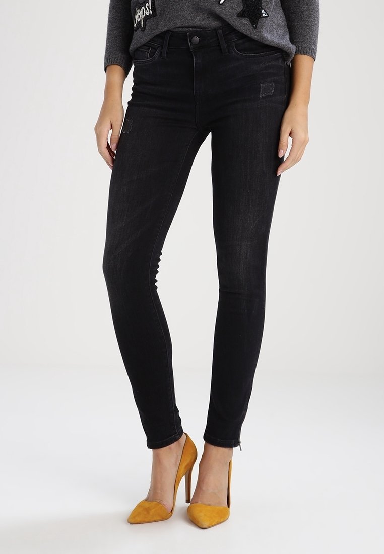 Jeans skinny noirs taille haute, avec des déchirures subtiles, jambe fuselée et design classique à cinq poches. Associés à des talons moutarde.