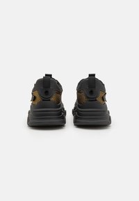 Zapatillas deportivas negras con suela acolchada, paneles laterales de malla en dorado y lengüetas en la parte trasera. Detalles texturizados en la suela.