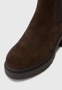 Ruskean mokkasaappaat nilkkapituisilla, pyöreällä varvalla, erottuvalla mustalla kumipohjalla ja joustavilla sivupaneeleilla. Sisältää tekstuuririkkaan ompelun yksityiskohtia.