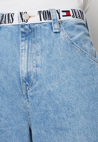 Gaiši zili džinsi ar platu jostasvietu, uz kuras ir melns "TOMMY JEANS" logotips un sarkans un balts emblemas akcenti.