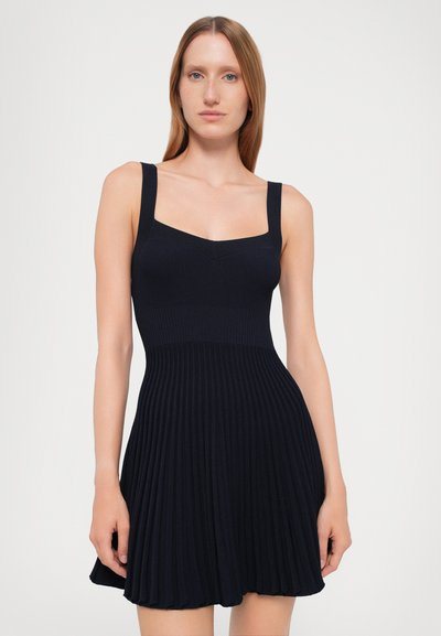 Navy geribbelde jurk met een fitted lijfje en een uitlopende plissé rok, voorzien van een vierkante halslijn en brede bandjes. Soepele textuur, boven de knie lengte.