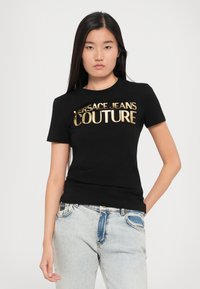 Melnas kokvilnas T-krekls ar zeltainu metāliski spīdīgu uzrakstu "VERSACE JEANS COUTURE". Pieguļošs dizains ar īsām piedurknēm un apaļu kakla izgriezumu.